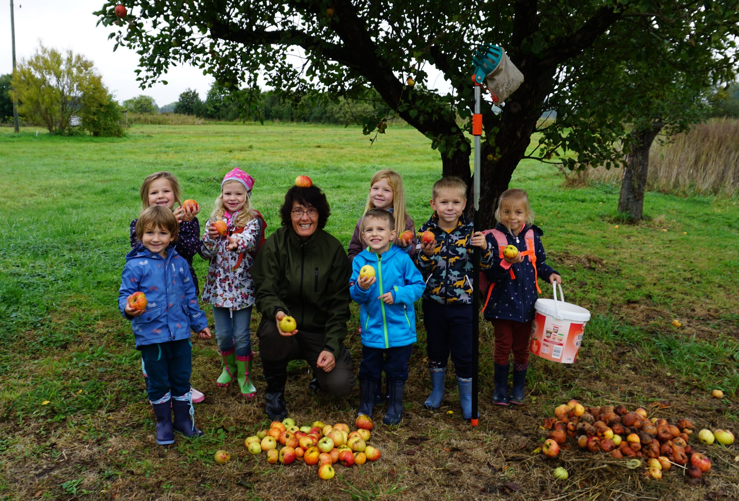 Mehrere Kinder mit einer Erzieherin stehen fröhlich mit Äpfeln in der Hand vor einem Apfelbaum